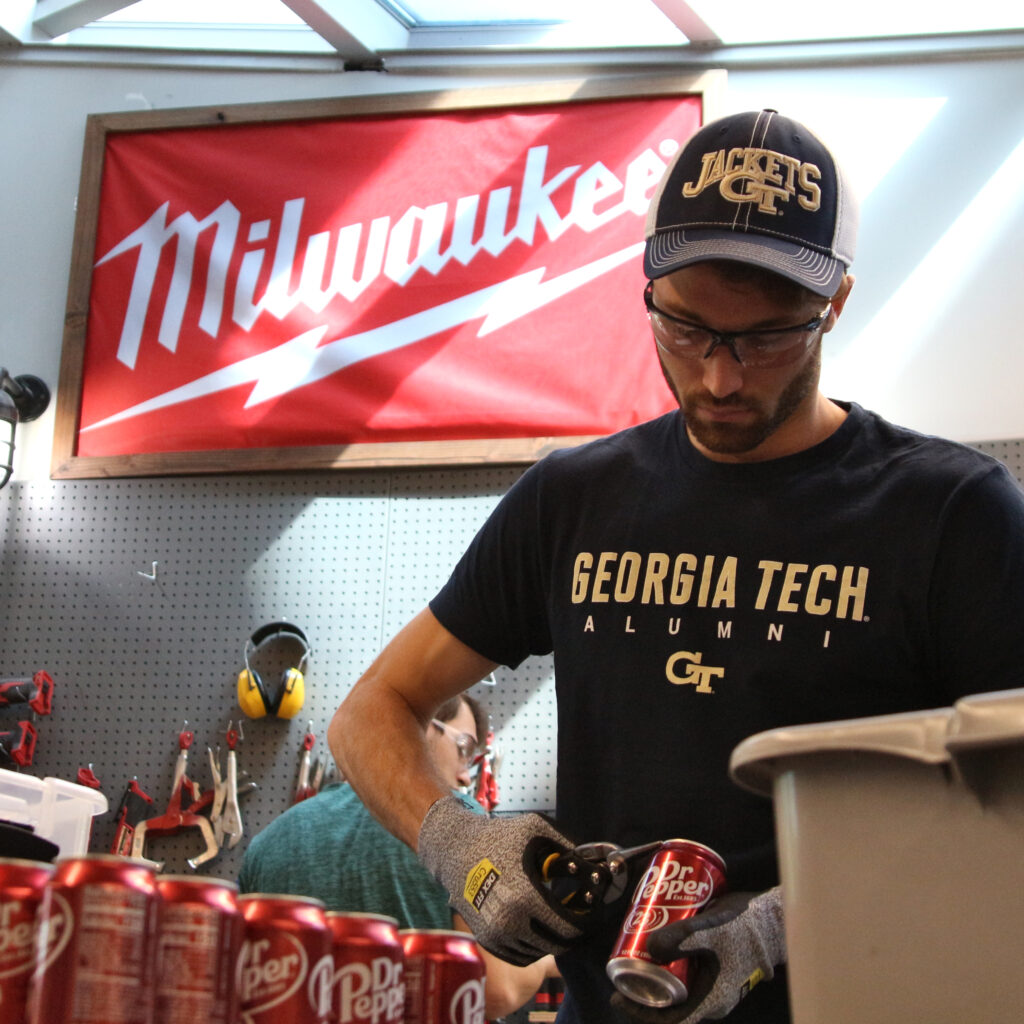 Photo of a person cutting up cans in the metal room with a Milwaukee flag behind him.